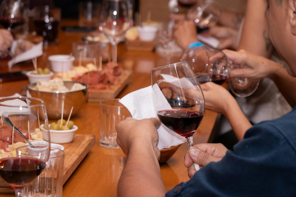 Close-up of people's hands holding wine glasses at a wine tasting event with snacks on a wooden table.